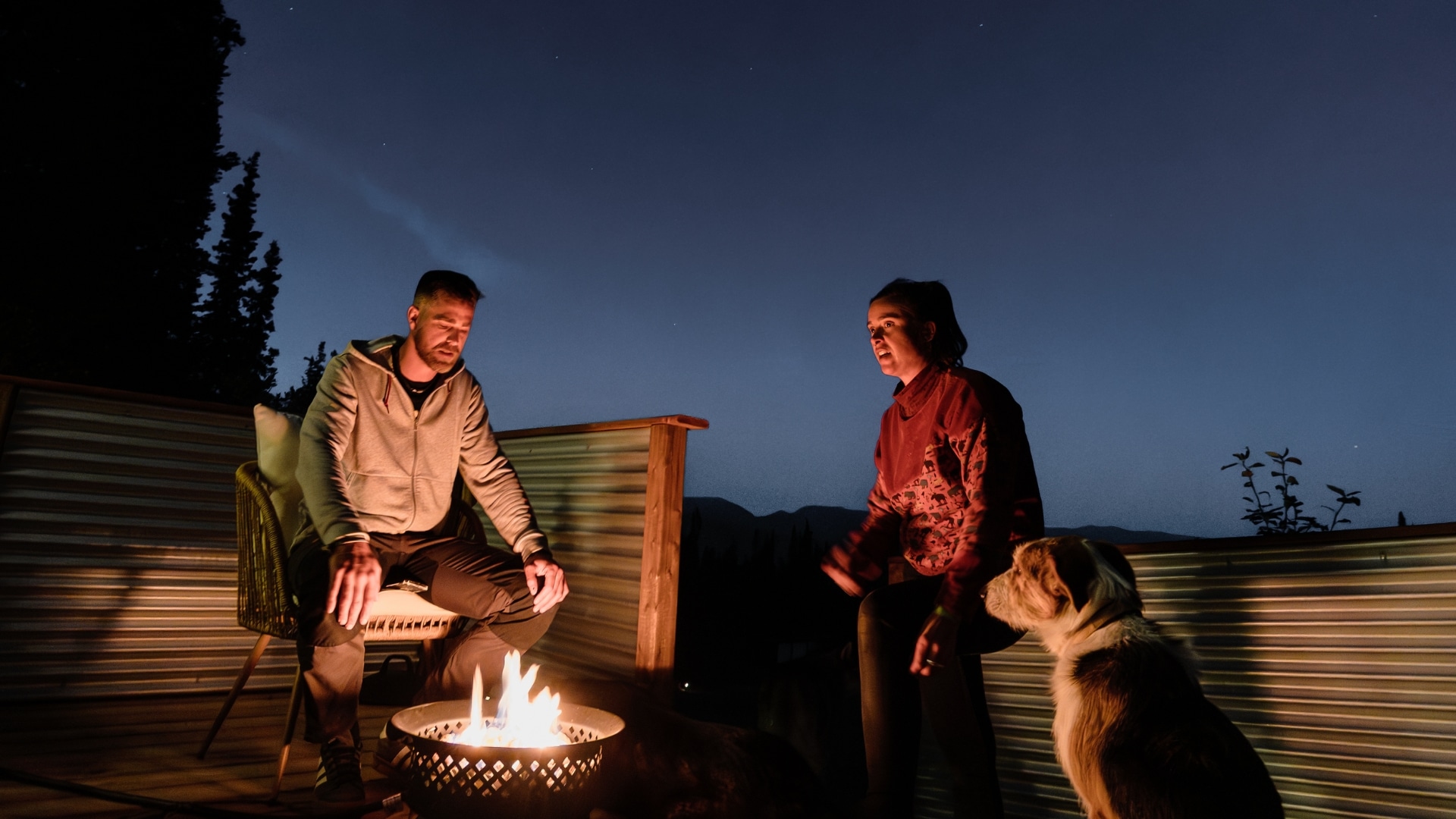 A young couple sit around a fire on a deck with the night sky behind them